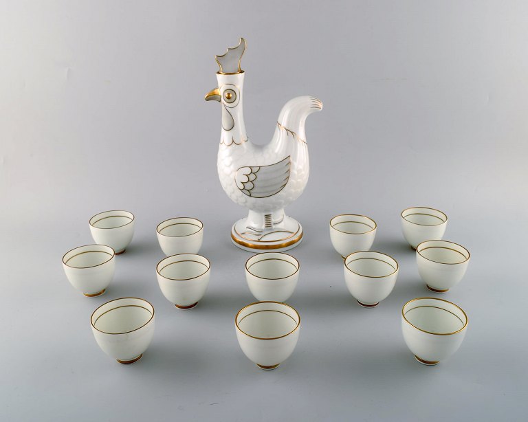 Bing & Grondahl/ B&G, Denmark. Jug of porcelain in the form of rooster, stopper with rooster comb. 12 accompanying porcelain mugs with gold-painted details.