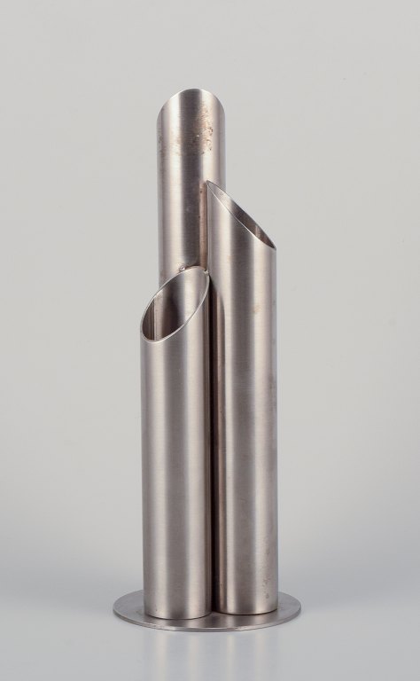 Svensk industriel designer. Vase i rustfrit stål udformet med tre sammensatte rør på rund sokkel.